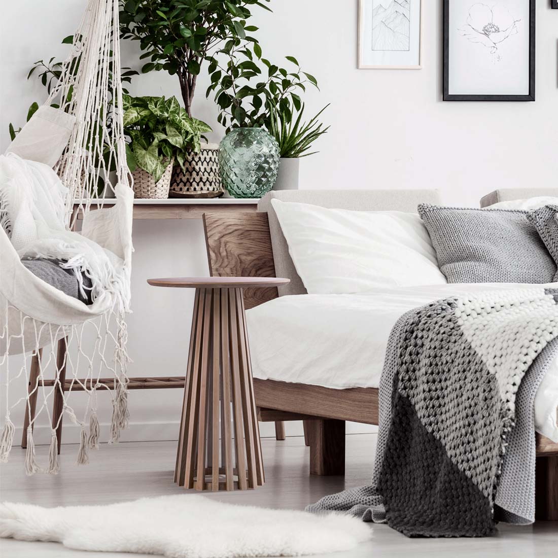 Load video: video showing a side table finished in black wood or natural oak wood, placed beside a bed.  Showing in is a large bed, with white bedding, grey pillows, grey throw blanket, many plants behind the bed on a table, and a macroma swing chair with many pillows in it.