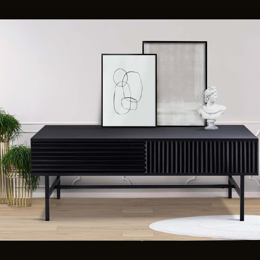 Load video: video showing a large black table in a wood finish that also has robust rion framework legs. Shown displayed in a foyer with plants to the left of it, and prints and a scuclputre ontop of the table